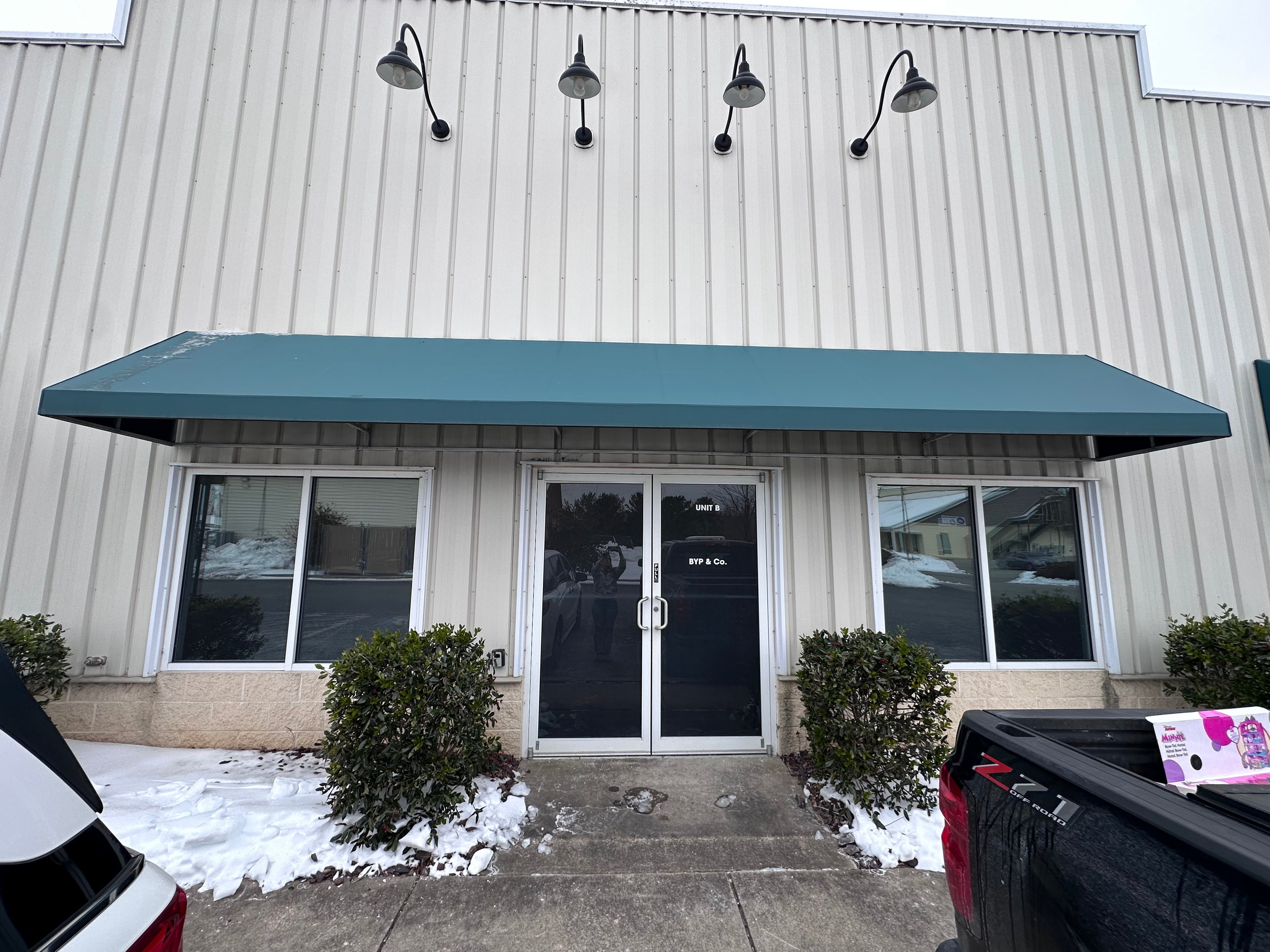 Commercial building entrance with a blue awning, snow on the ground, and vehicles parked.